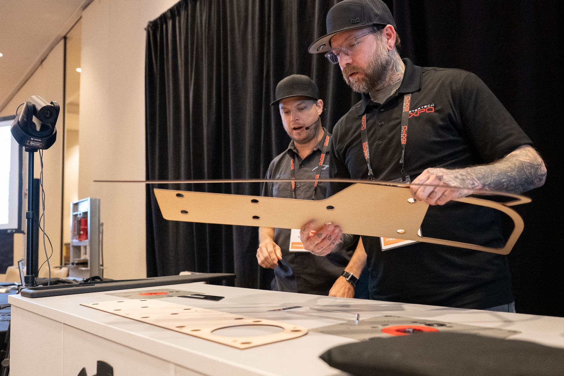 Two men examining a large, cut-out piece of tan material at a display table. One holds it up, the other looks on.