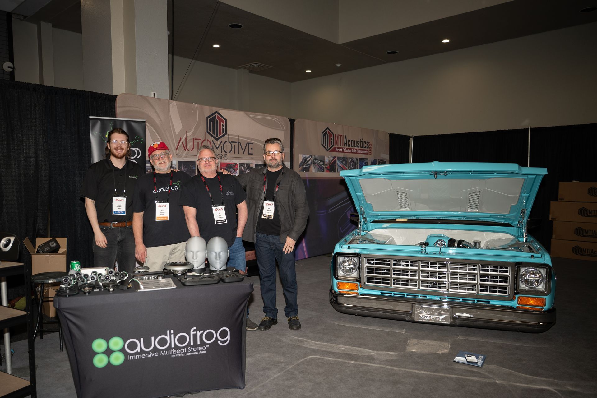 Four men standing near a turquoise truck with open hood; display table with 