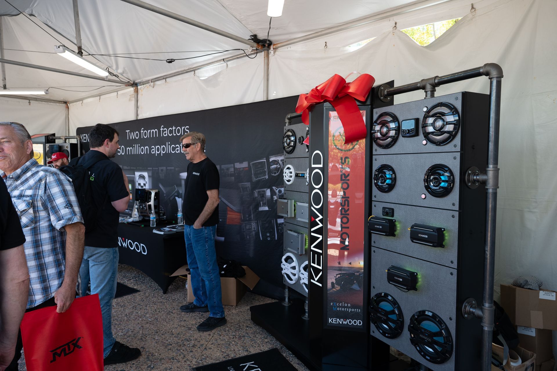Kenwood audio equipment display at an outdoor event. People browse products in a tent with black backdrop.