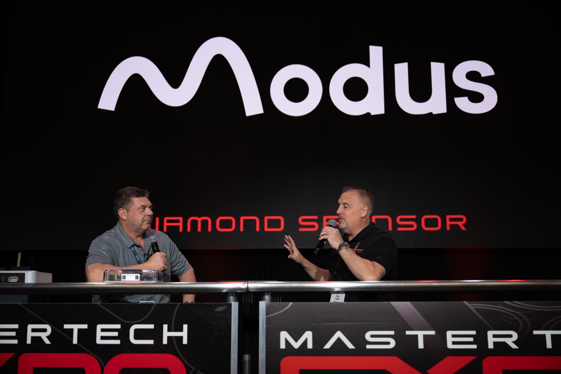 Two men on stage with a Modus logo backdrop; one speaks into a microphone, the other listens.