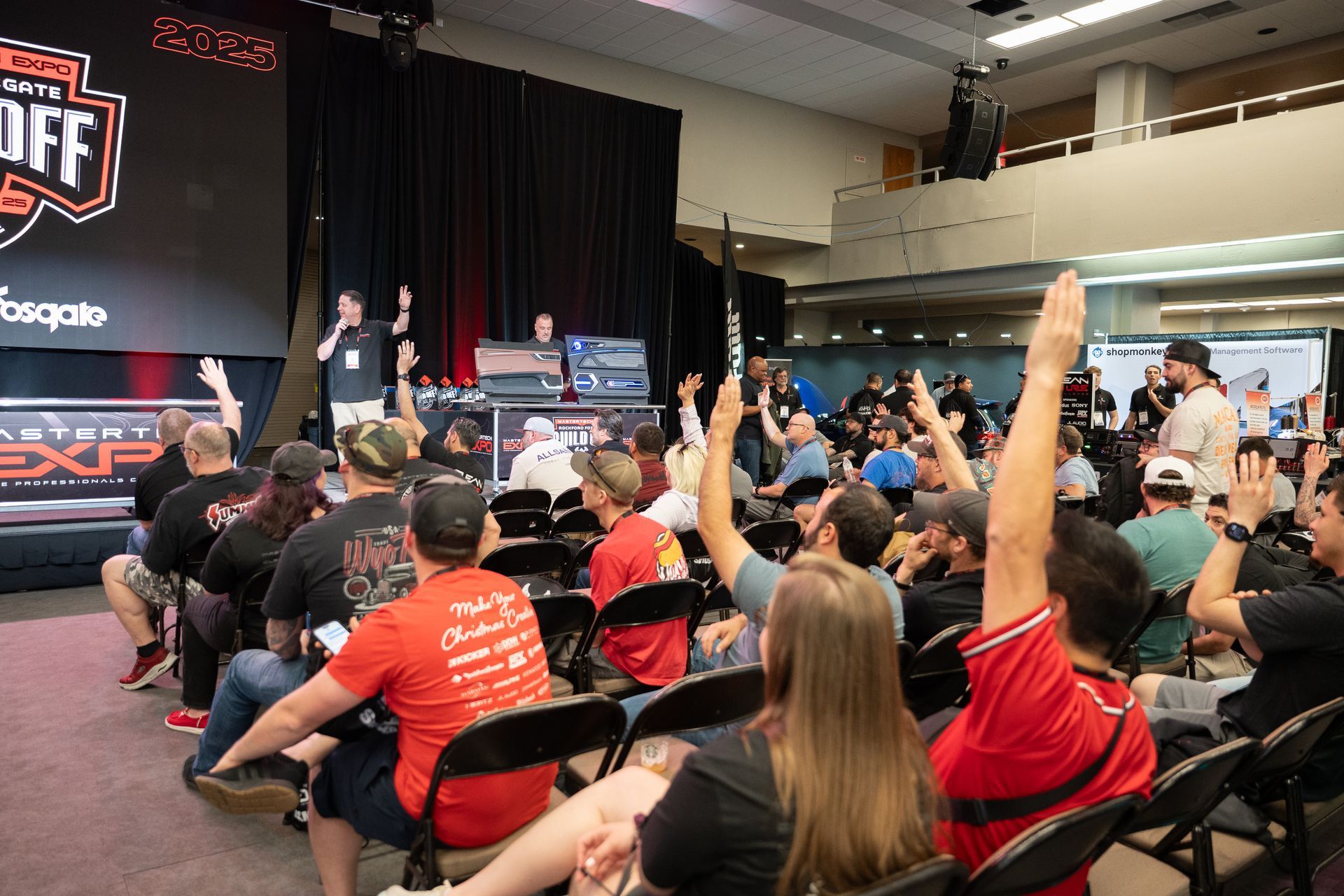 A large crowd at an event, many with hands raised, presumably bidding. Auctioneer on stage.