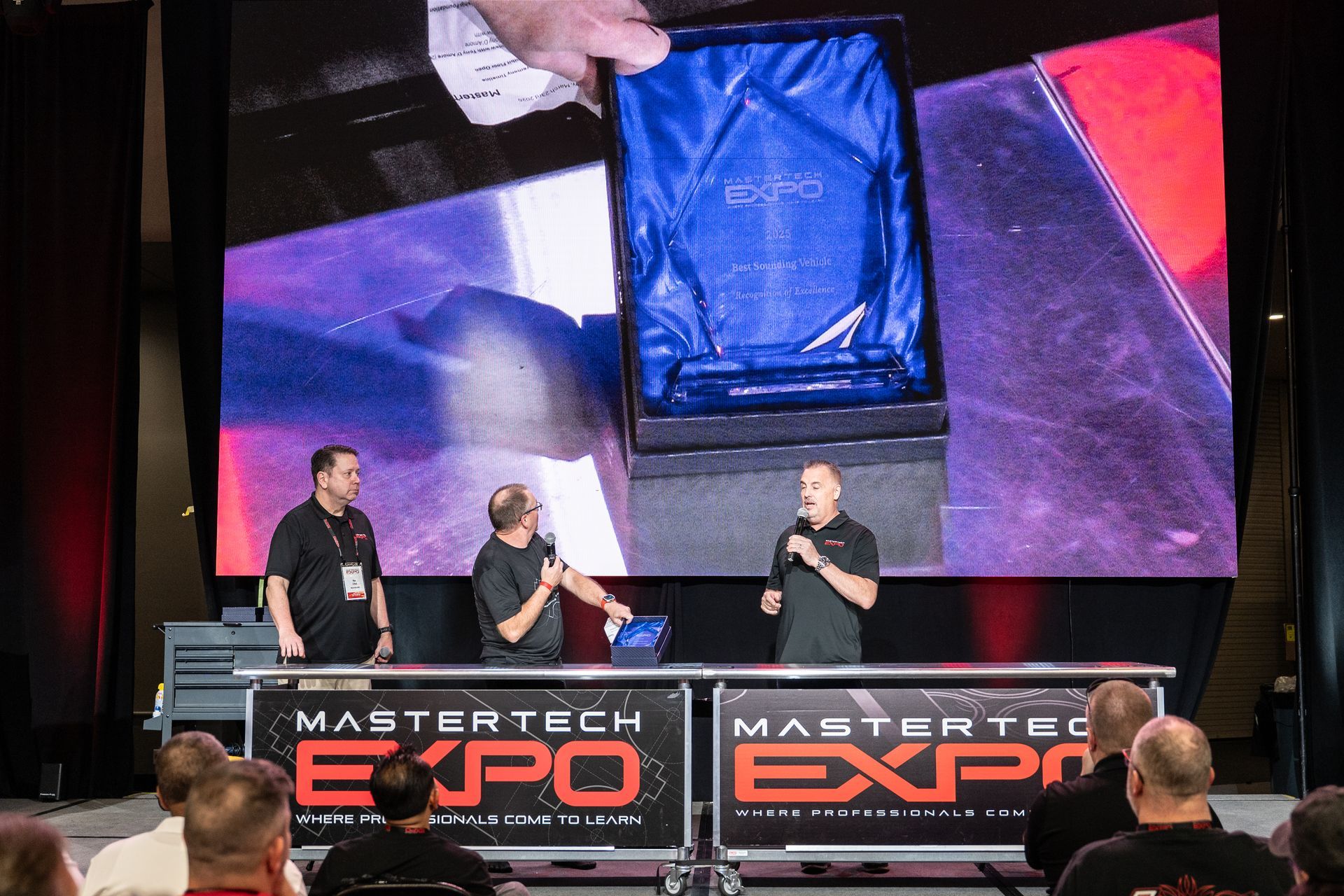 Men presenting an award on stage at the Mastertech Expo. A screen displays the event's logo.