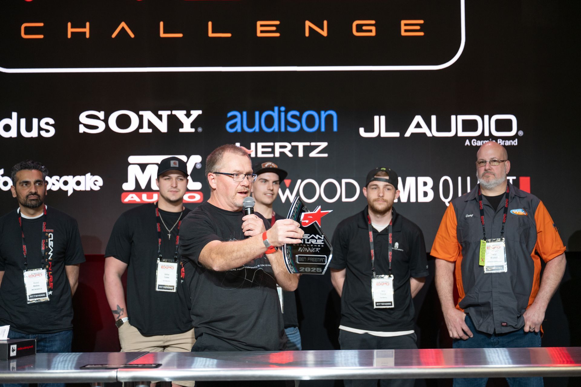 Group of men at an event, one holding a trophy, sponsors logos in the background.