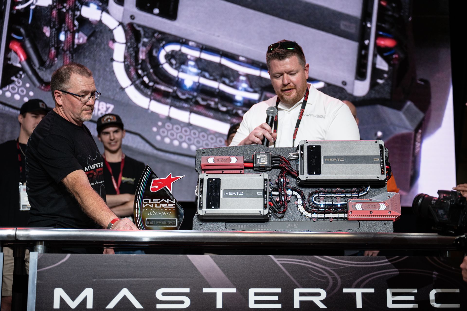 Two men at MasterTech event, one speaking, examining electronics display. Trophy and onlookers visible.