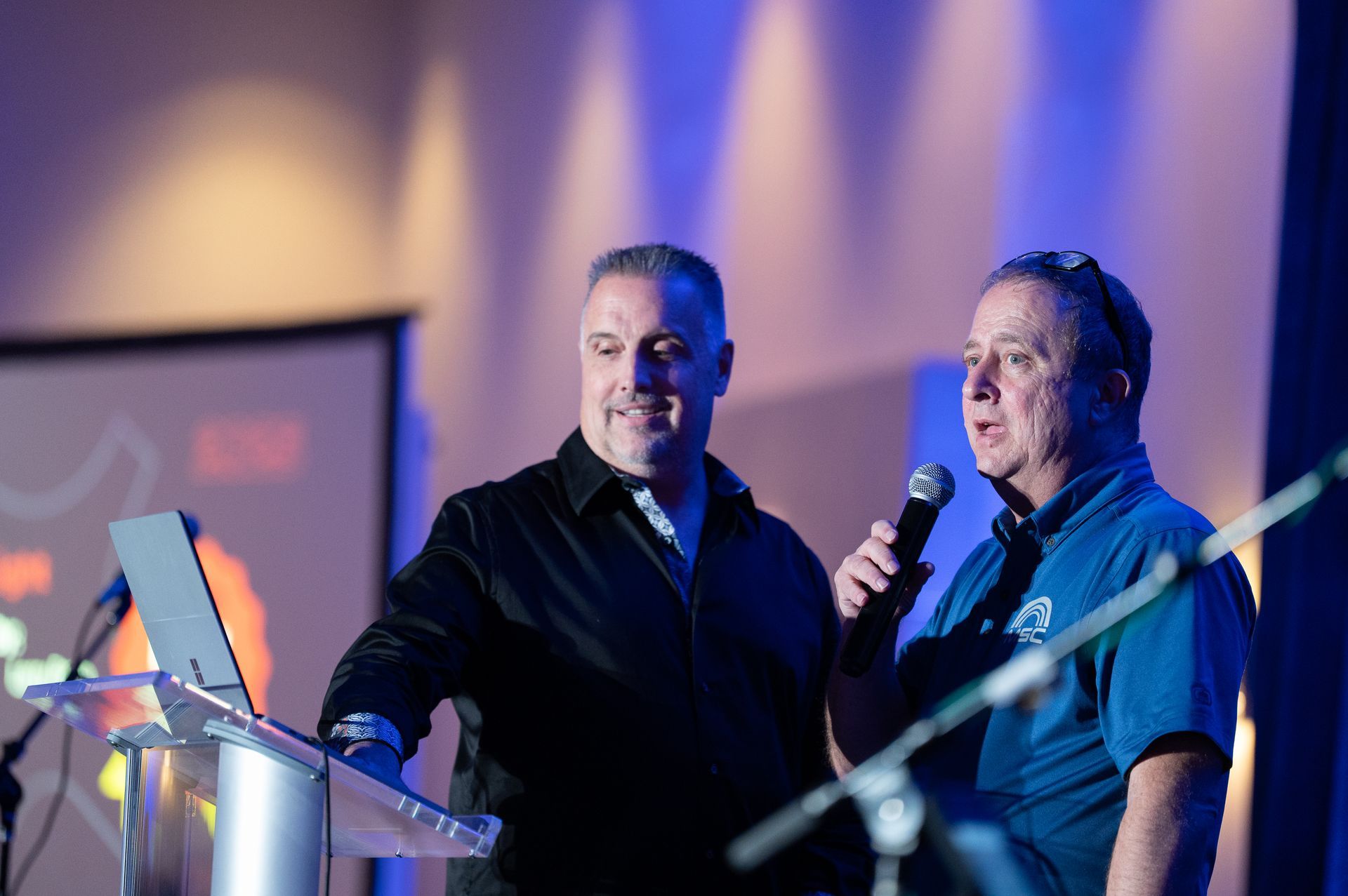 Two men at a podium; one speaks into a microphone, the other stands next to a laptop; stage setting.