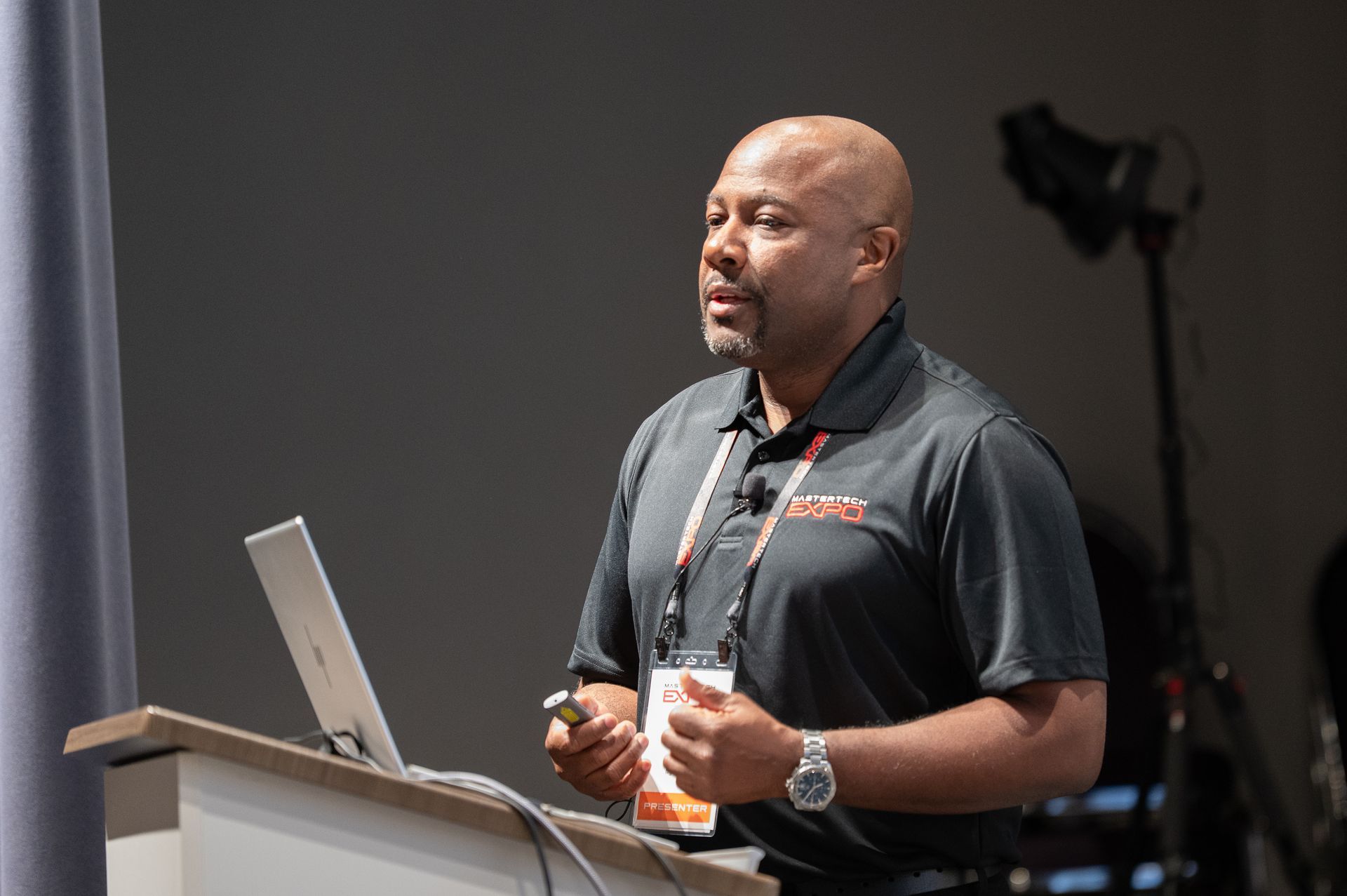 Man giving a presentation at a podium, wearing a dark shirt and holding a clicker.
