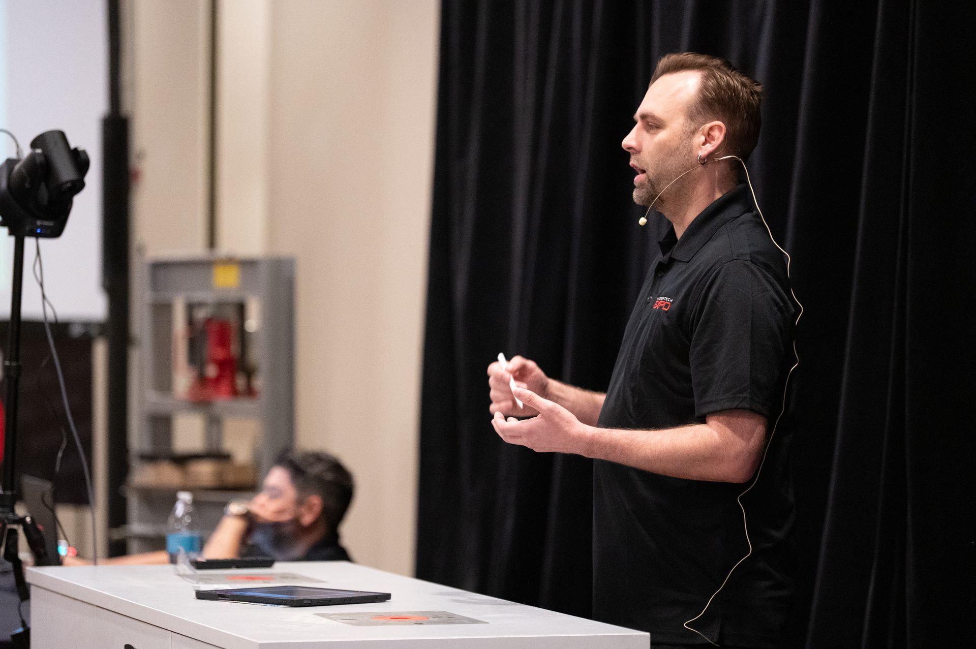 Man giving a presentation, holding a marker. A person and equipment visible in background.