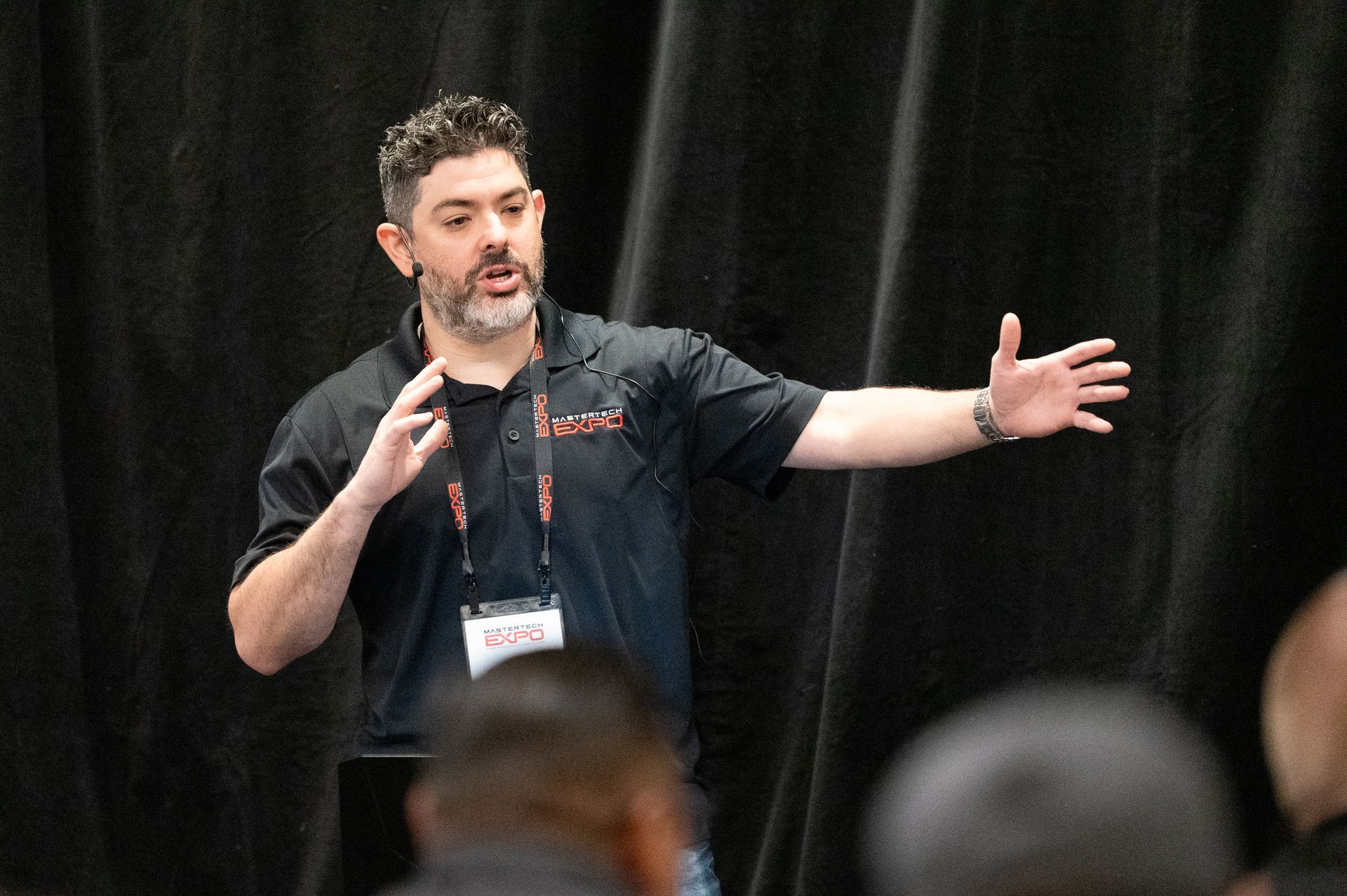 Man gesturing with hands while speaking to an audience, wearing a black shirt with a lanyard.