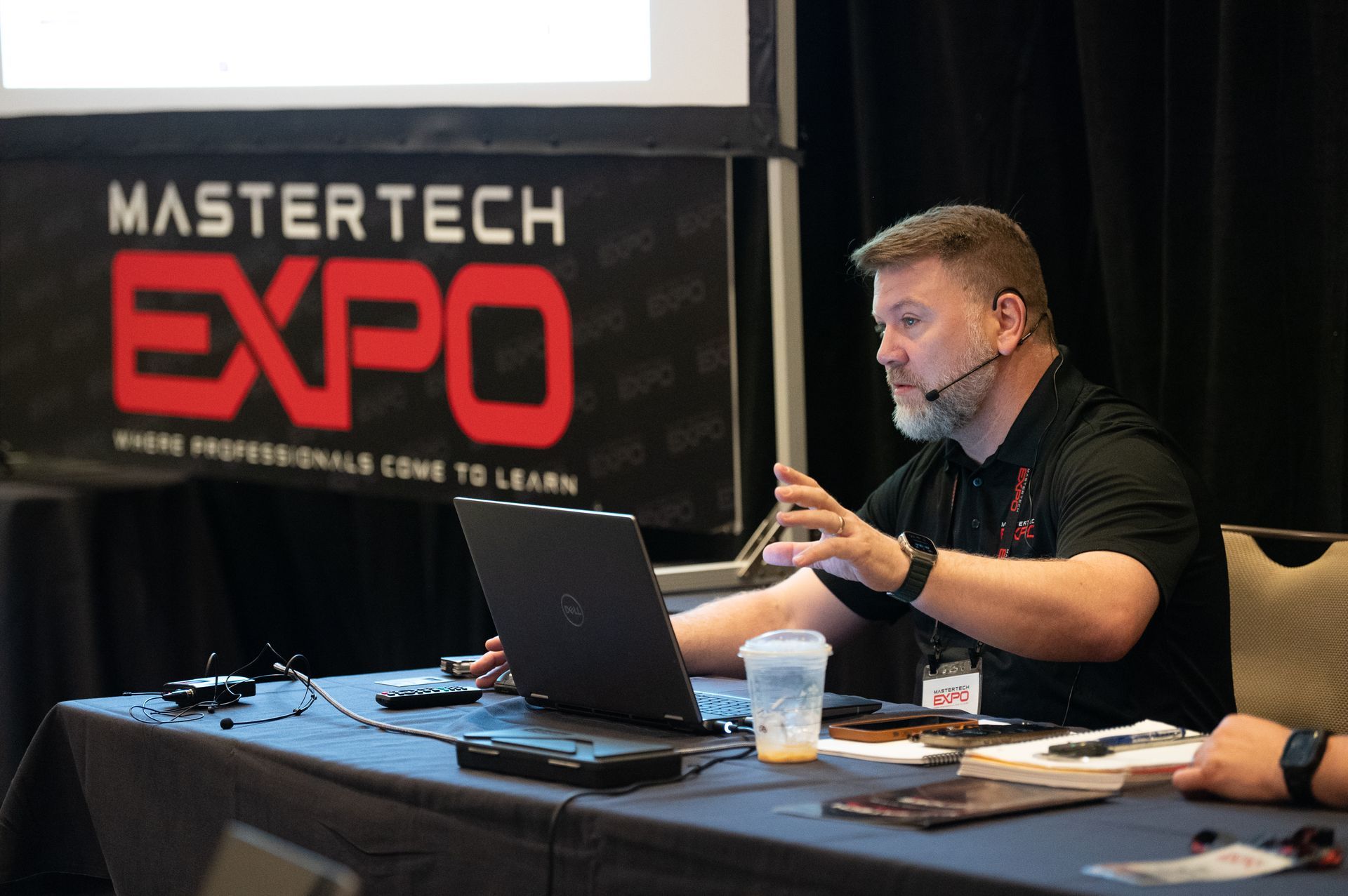 Man presenting at Mastertech Expo, gesturing with hands, laptop on table. Expo logo visible.