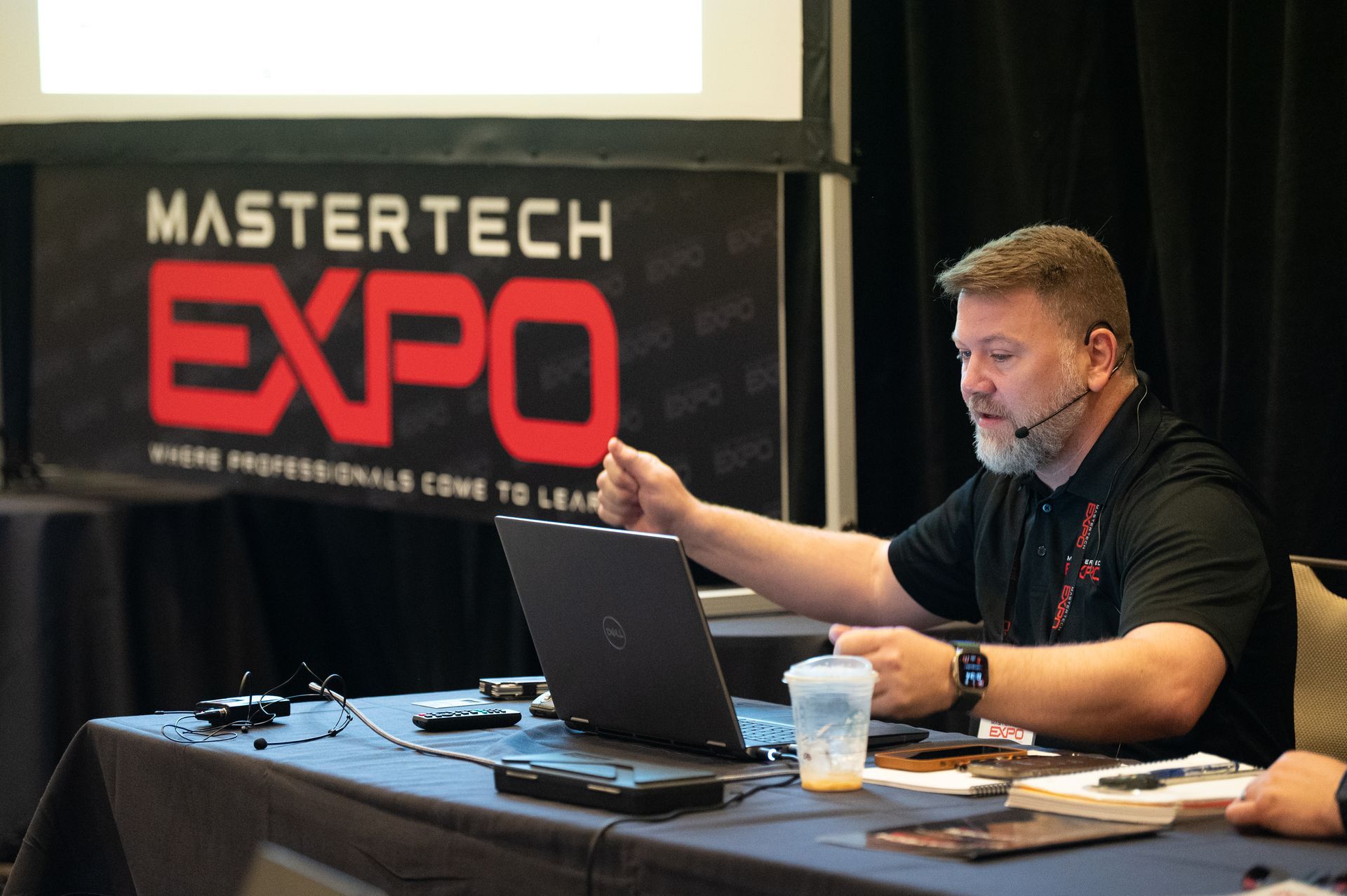Man presenting at the Mastertech Expo, gesturing towards laptop, microphone, water cup, banner in background.