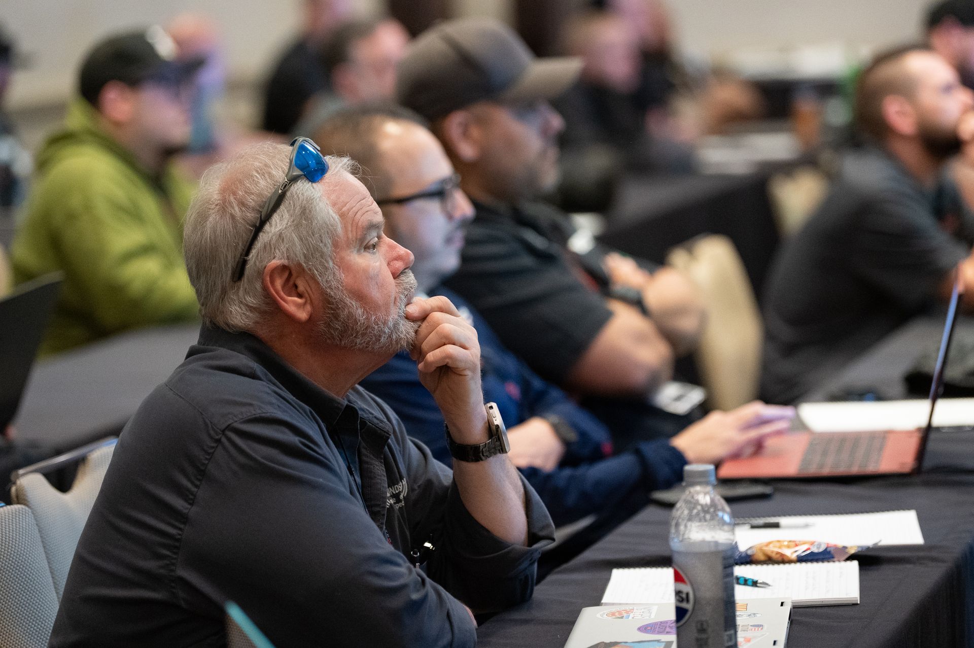 Attendees at a conference attentively watch a presentation. Man in front with glasses and gray beard.
