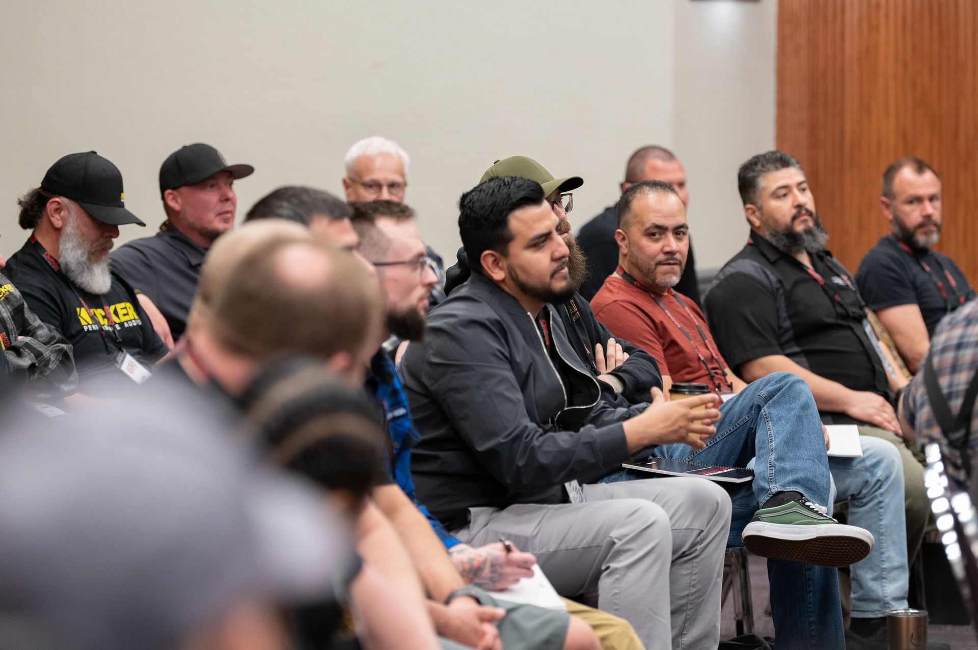 Audience listening in a conference room; several men in casual attire, some with beards, looking toward the speaker.