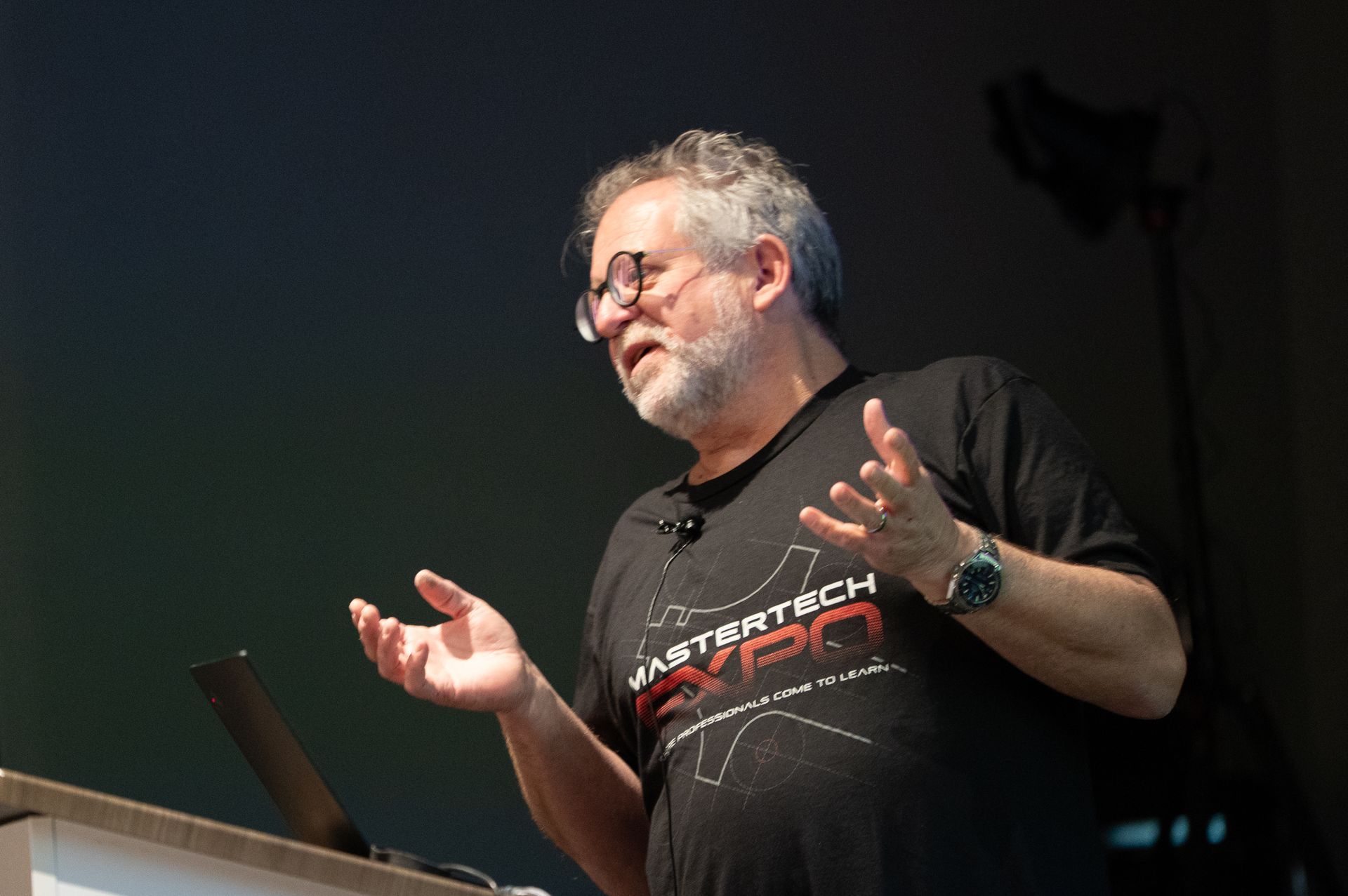 Man speaking at a conference, gesturing with hands. He's wearing a t-shirt, glasses, and a watch. Laptop on the podium.