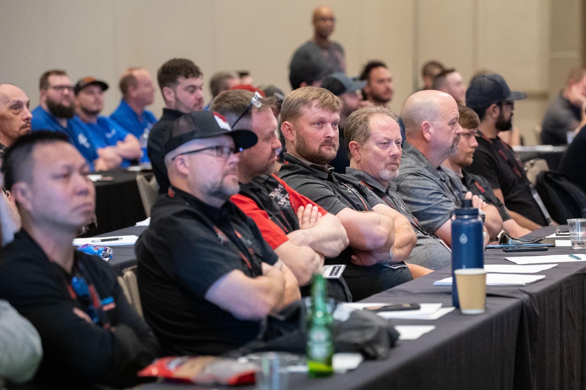 Audience in a conference room; men seated at tables, attentively listening to a presentation.