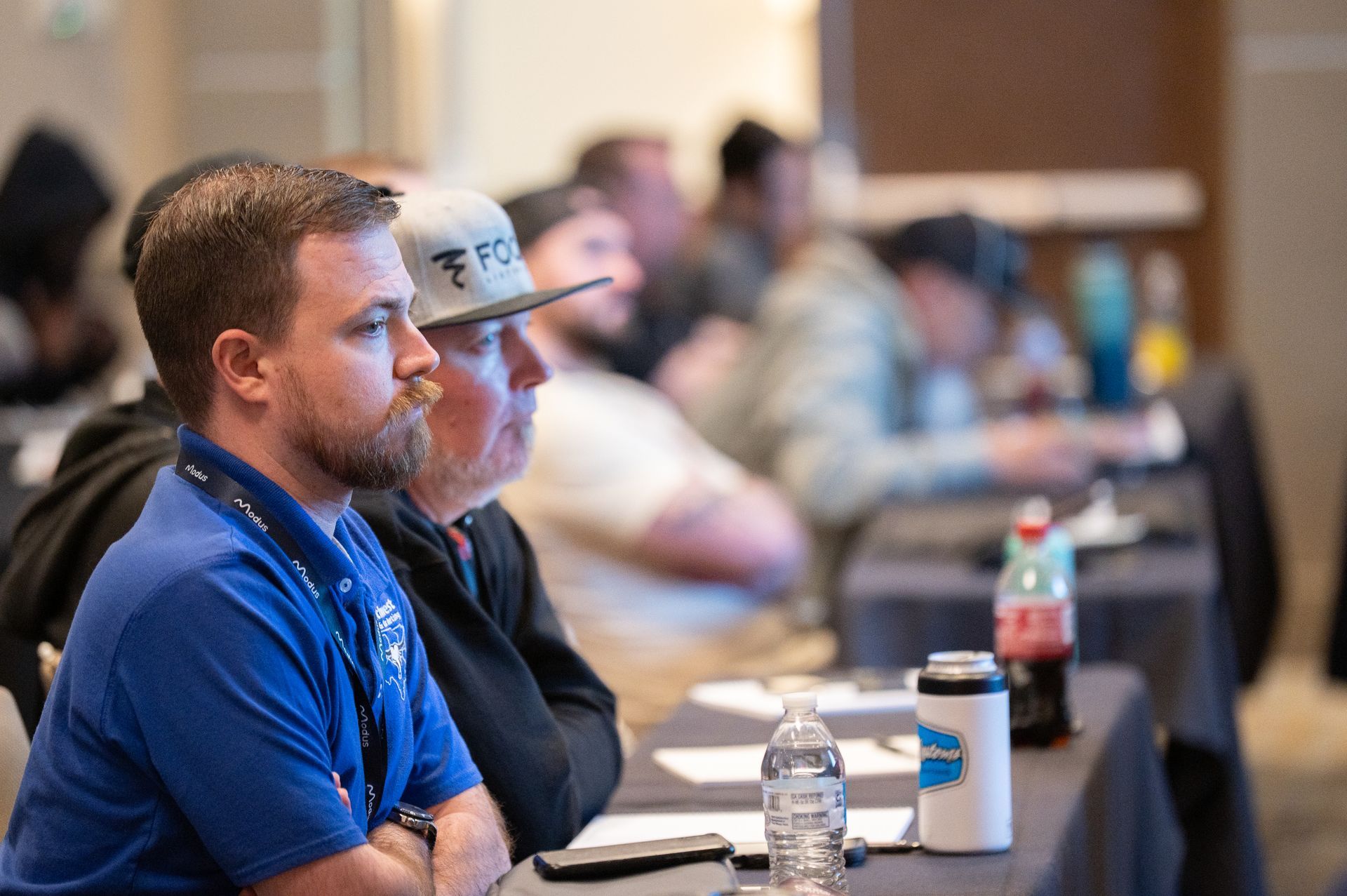 Men in a conference, listening intently. Some are wearing hats, drinks on the table.