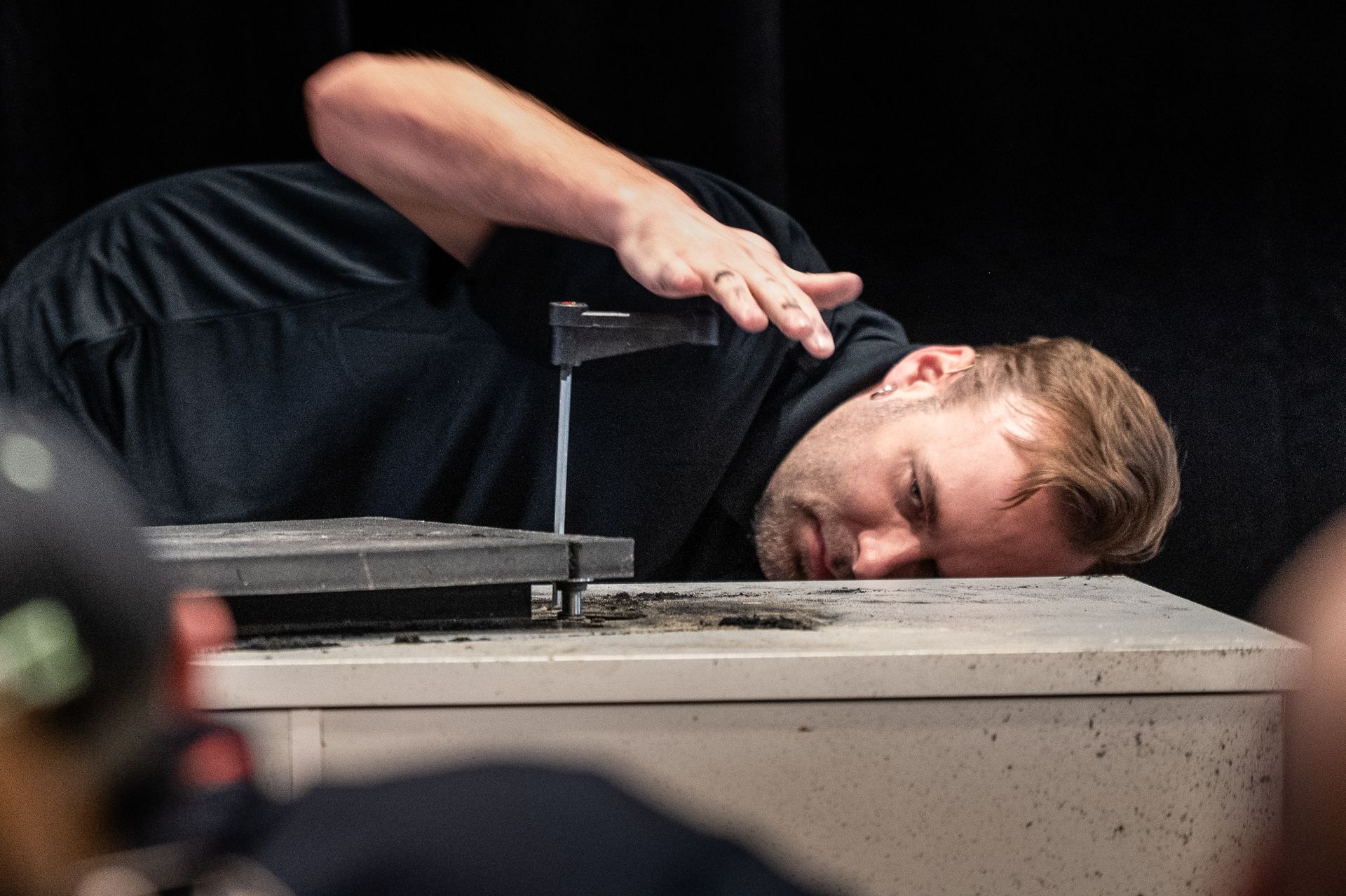 Man pouring liquid from a tool onto a table, eyes focused. Dark setting.