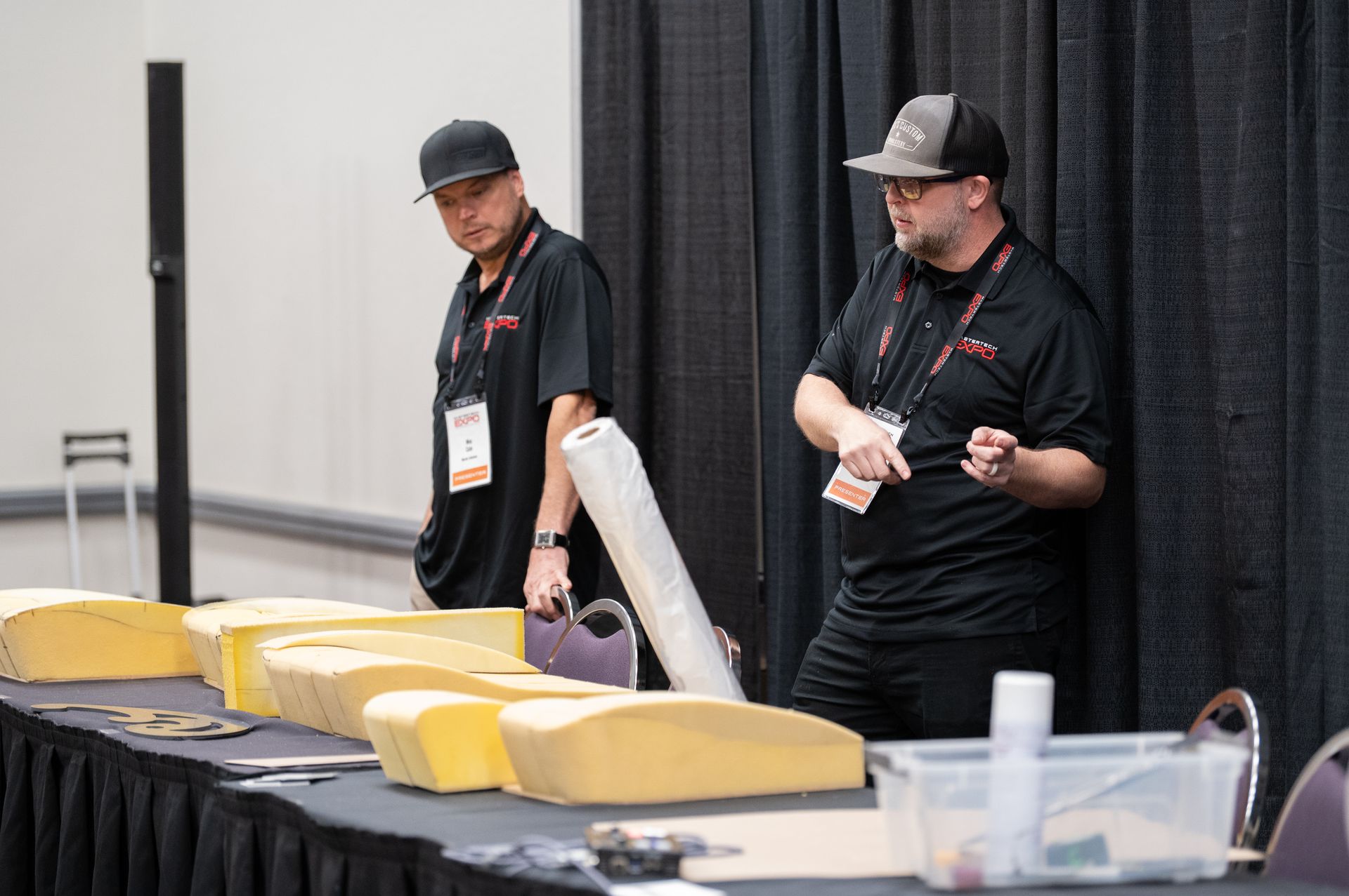Two men at a table with foam samples, possibly a trade show. One man gestures while talking, the other looks down.