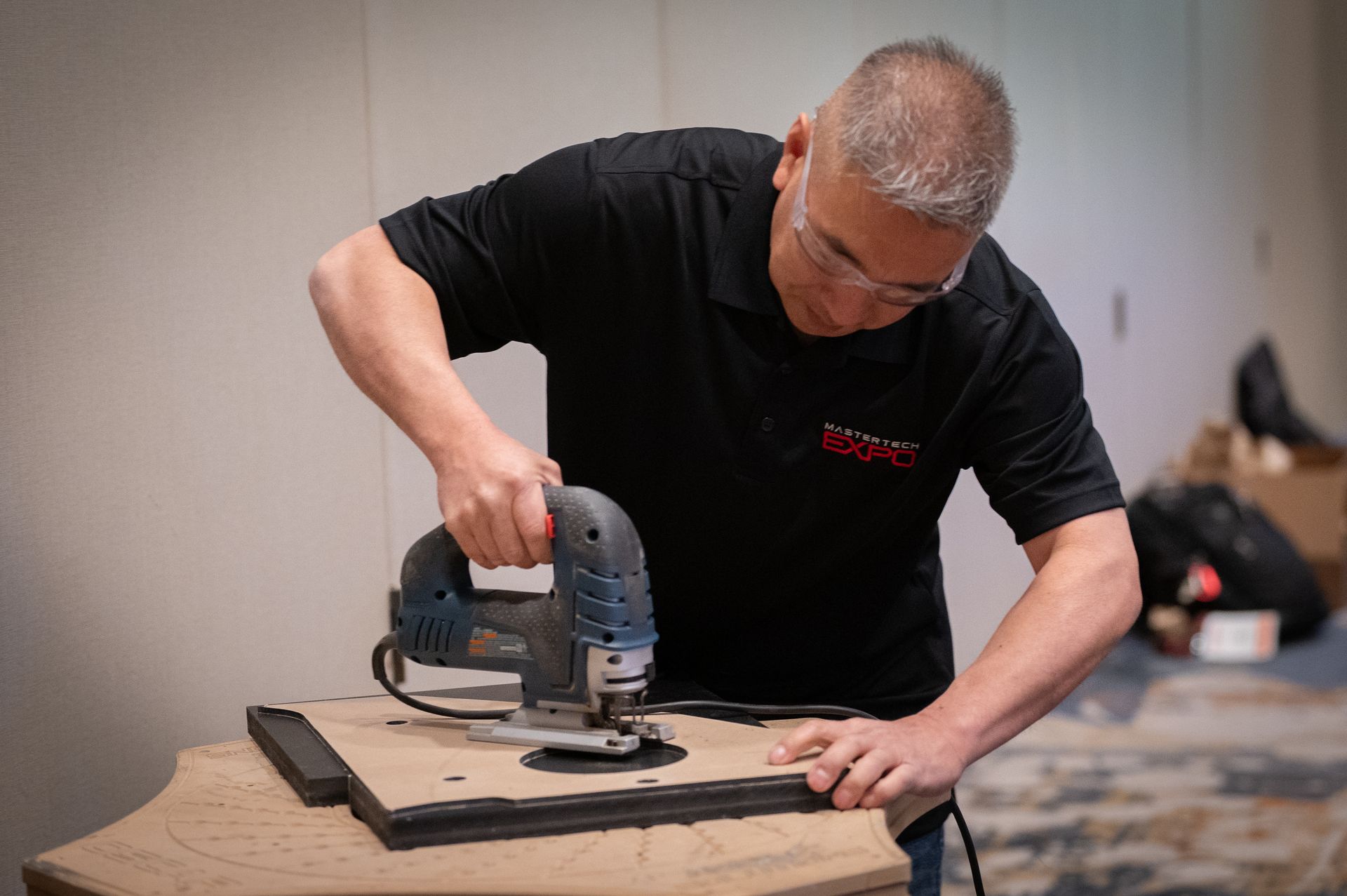 Man using a jigsaw to cut a piece of wood, wearing safety glasses and a black shirt.
