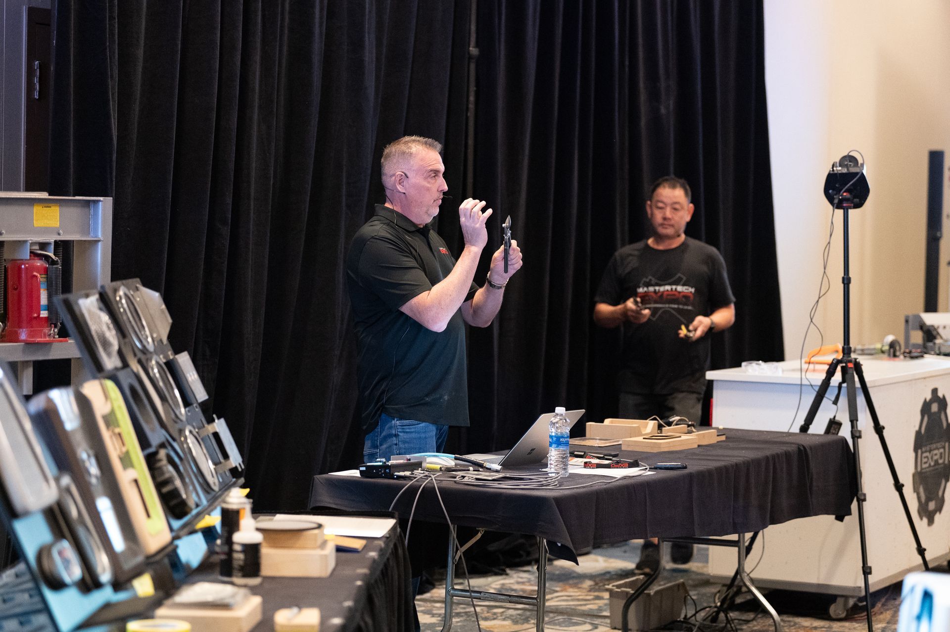 Man demonstrating tools at a table. Another man stands behind him. Tools and equipment on display.