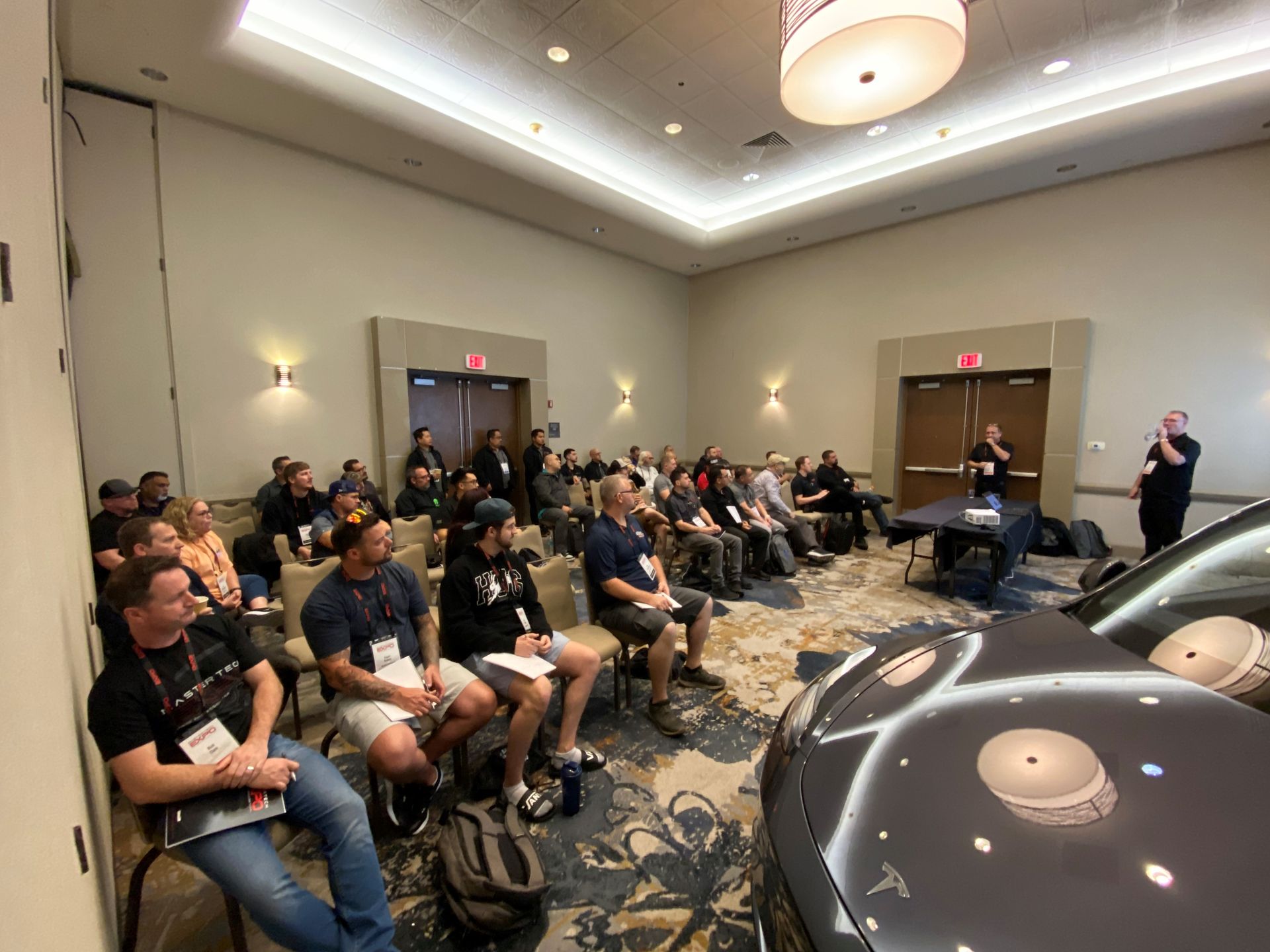 A group of people are sitting in a room with a car in the background.
