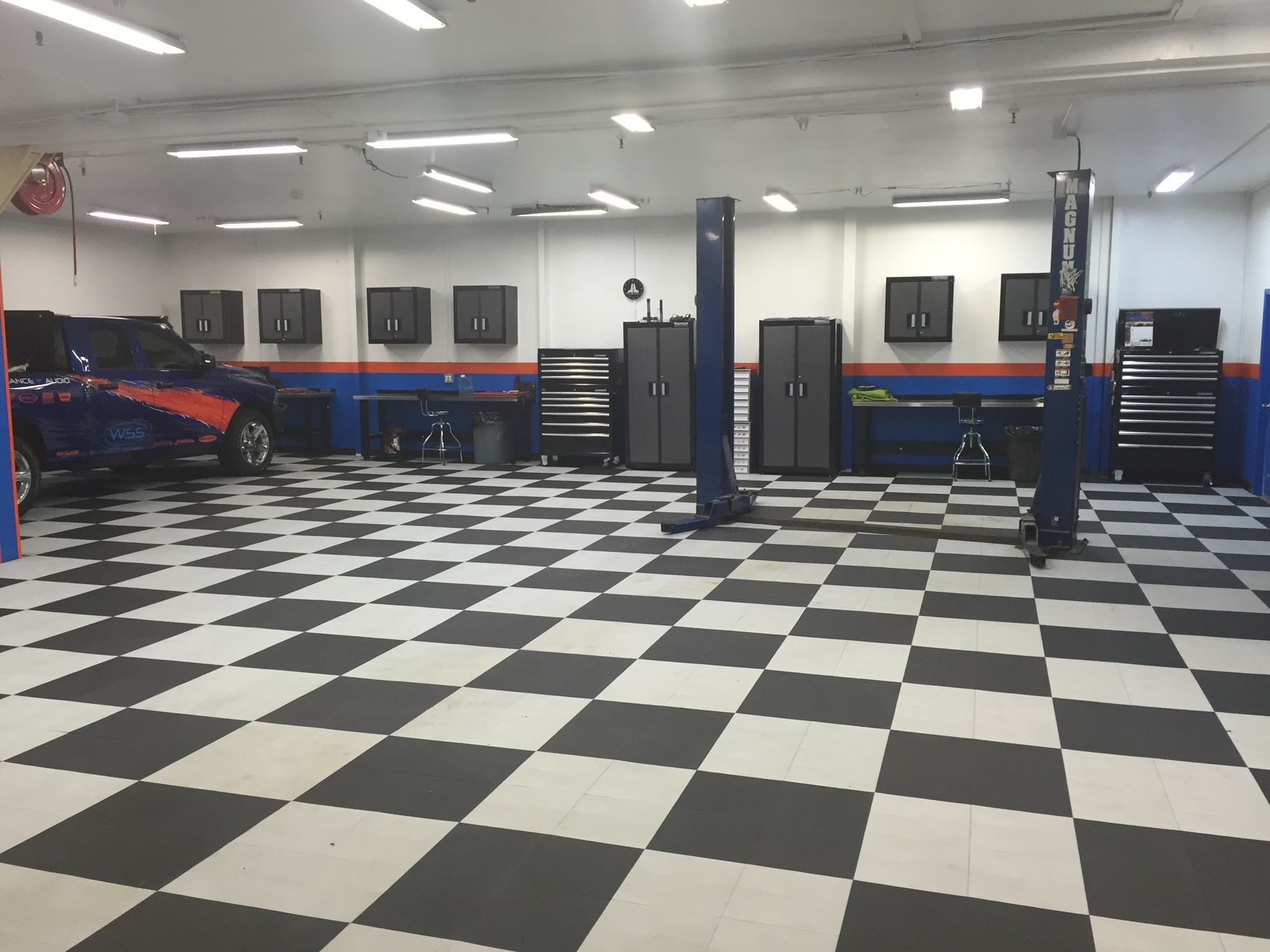 Garage interior with checkered floor, car, lifts, cabinets, and workbenches.