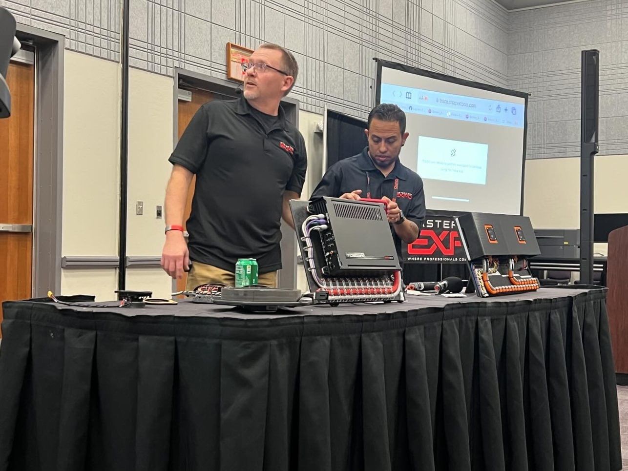 Two men at a presentation with audio equipment. One man looks towards the audience. The other works on the device.