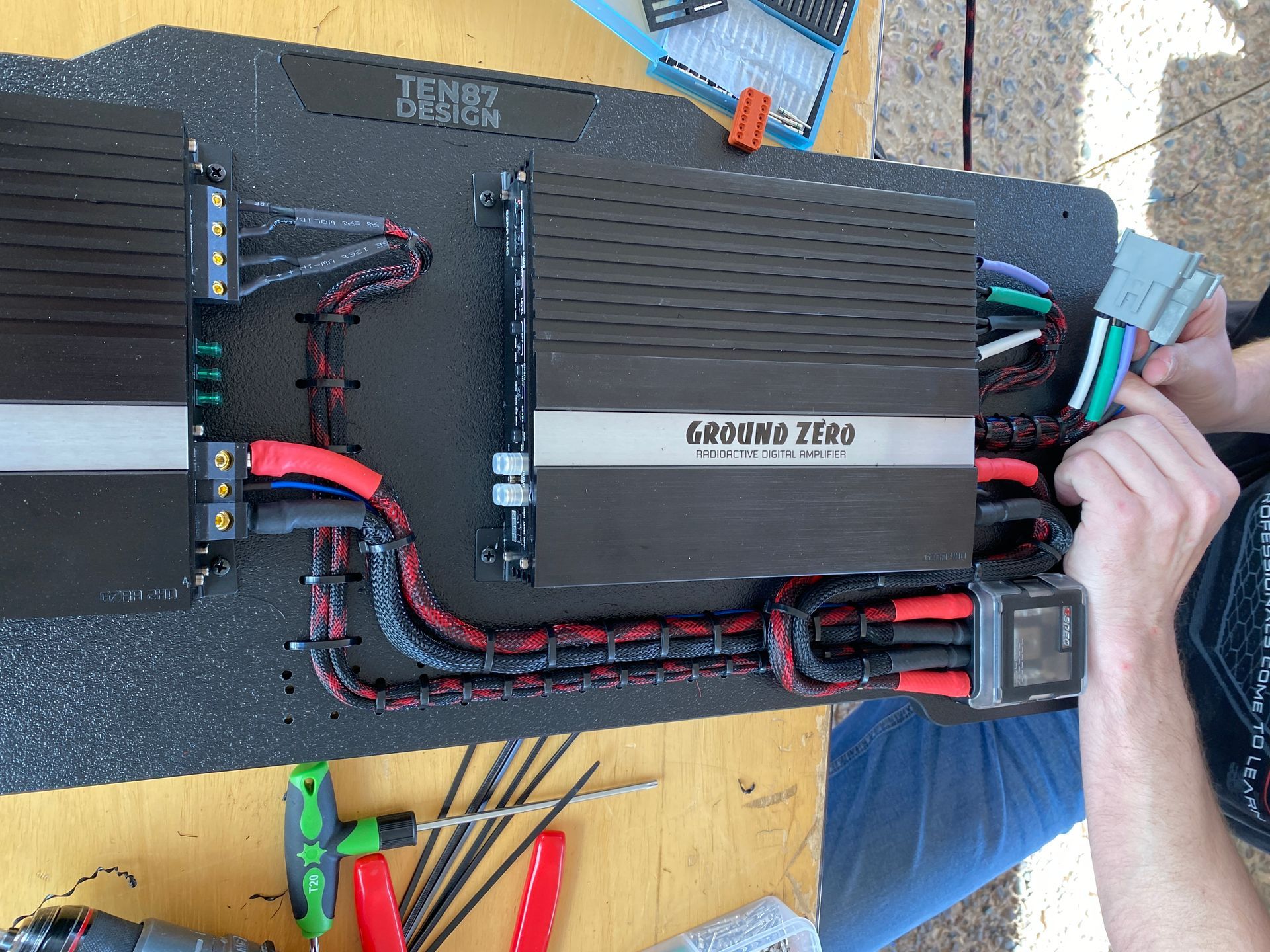 Person wiring car audio amplifiers on a black board. Red and black wires with tools scattered around.