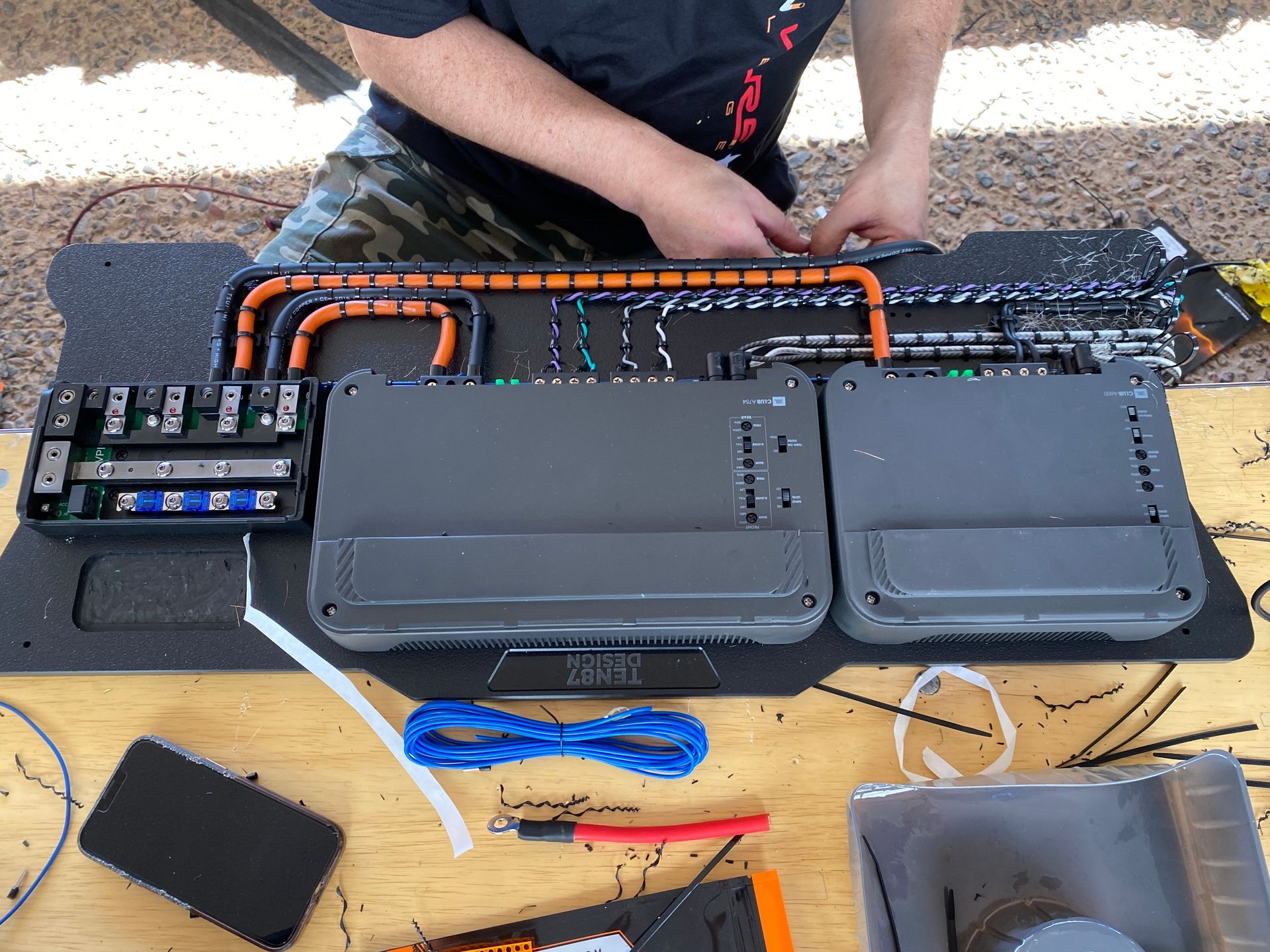 Person wiring car audio amplifiers on a wooden table outdoors. Wires, amps, and other equipment visible.