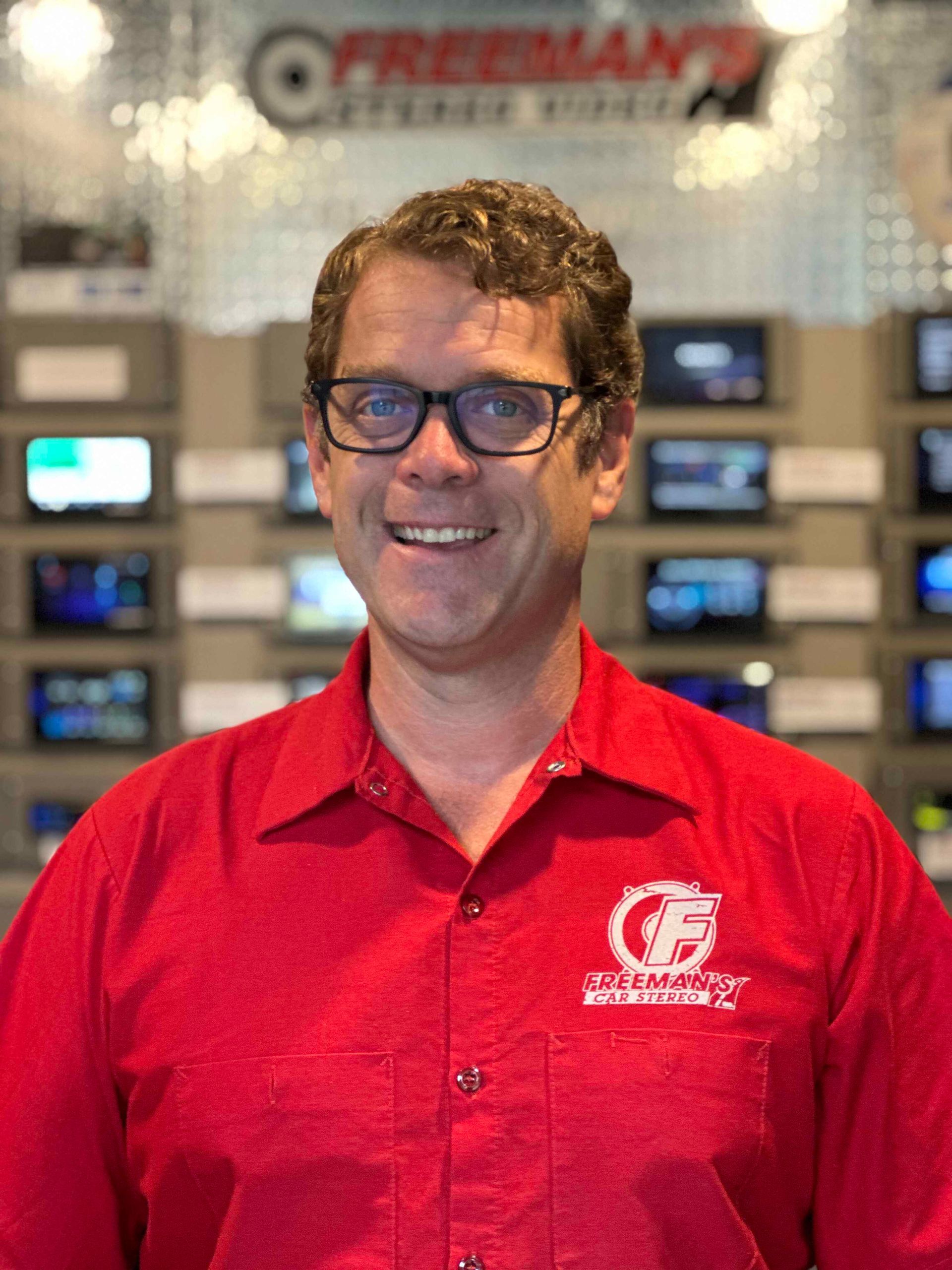 A man wearing glasses and a red shirt is standing in front of a wall of televisions.