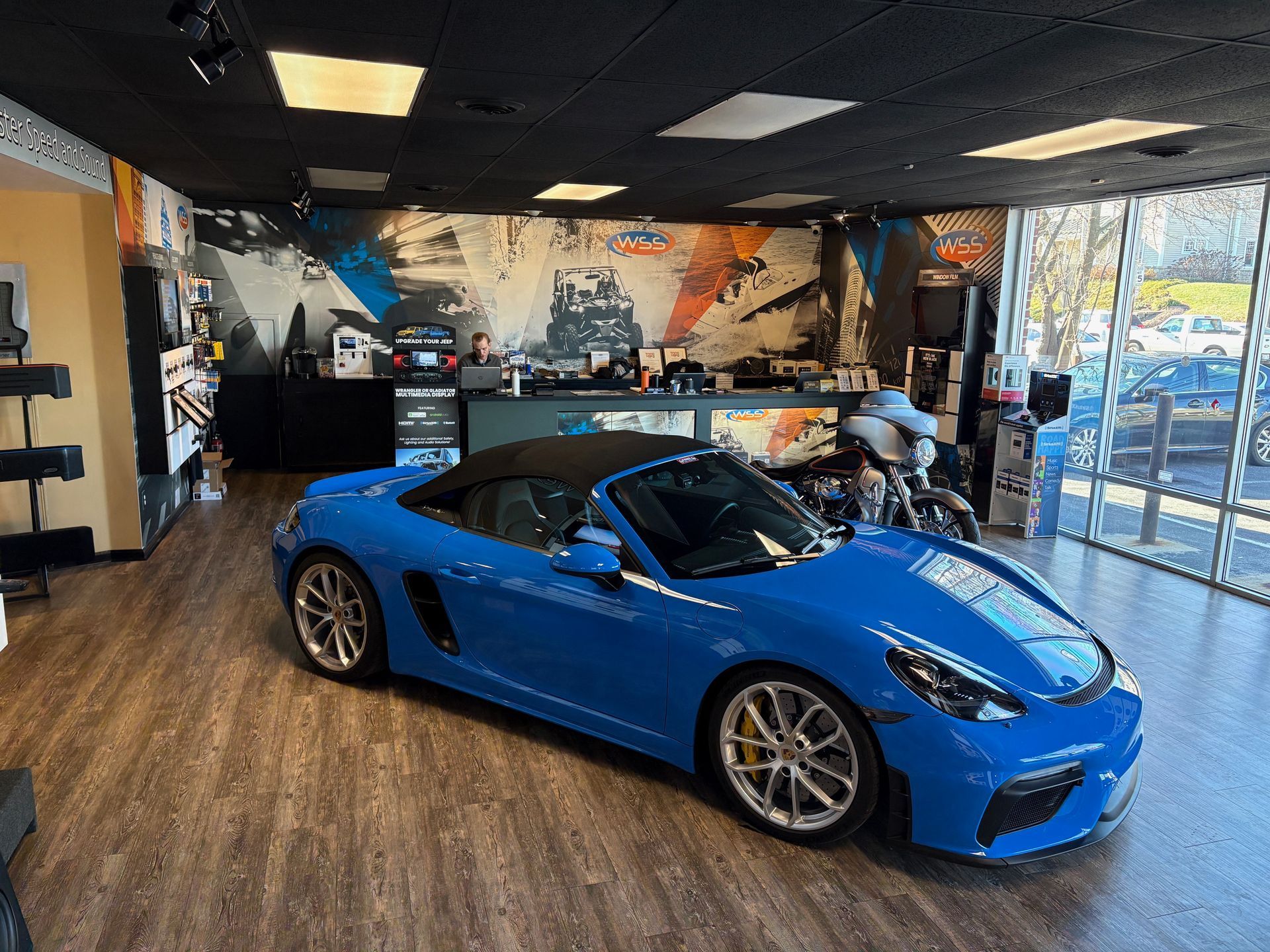 Blue convertible Porsche in a showroom with car art and window view.