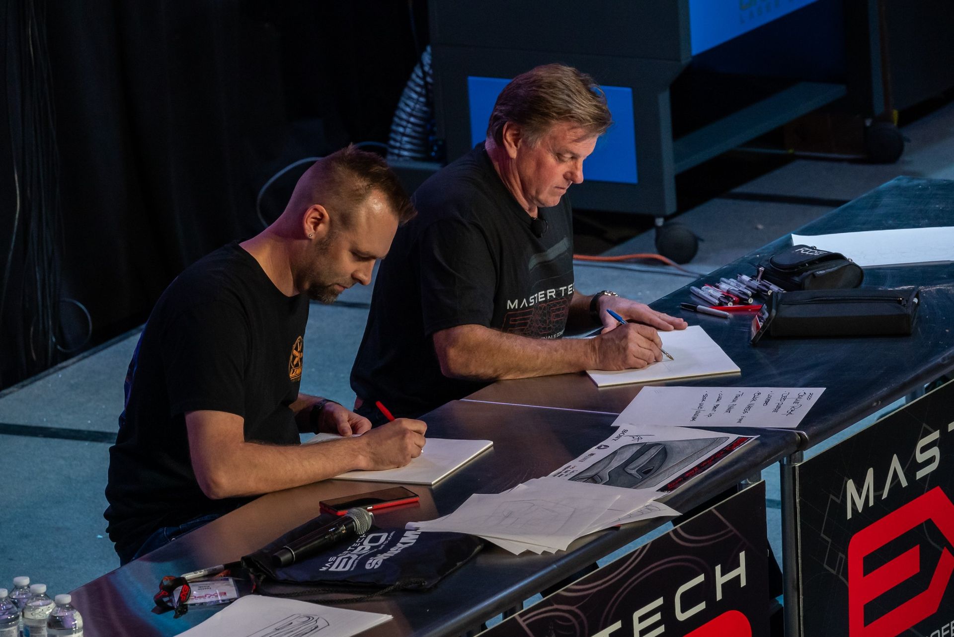 Two men are sitting at a table writing on papers.