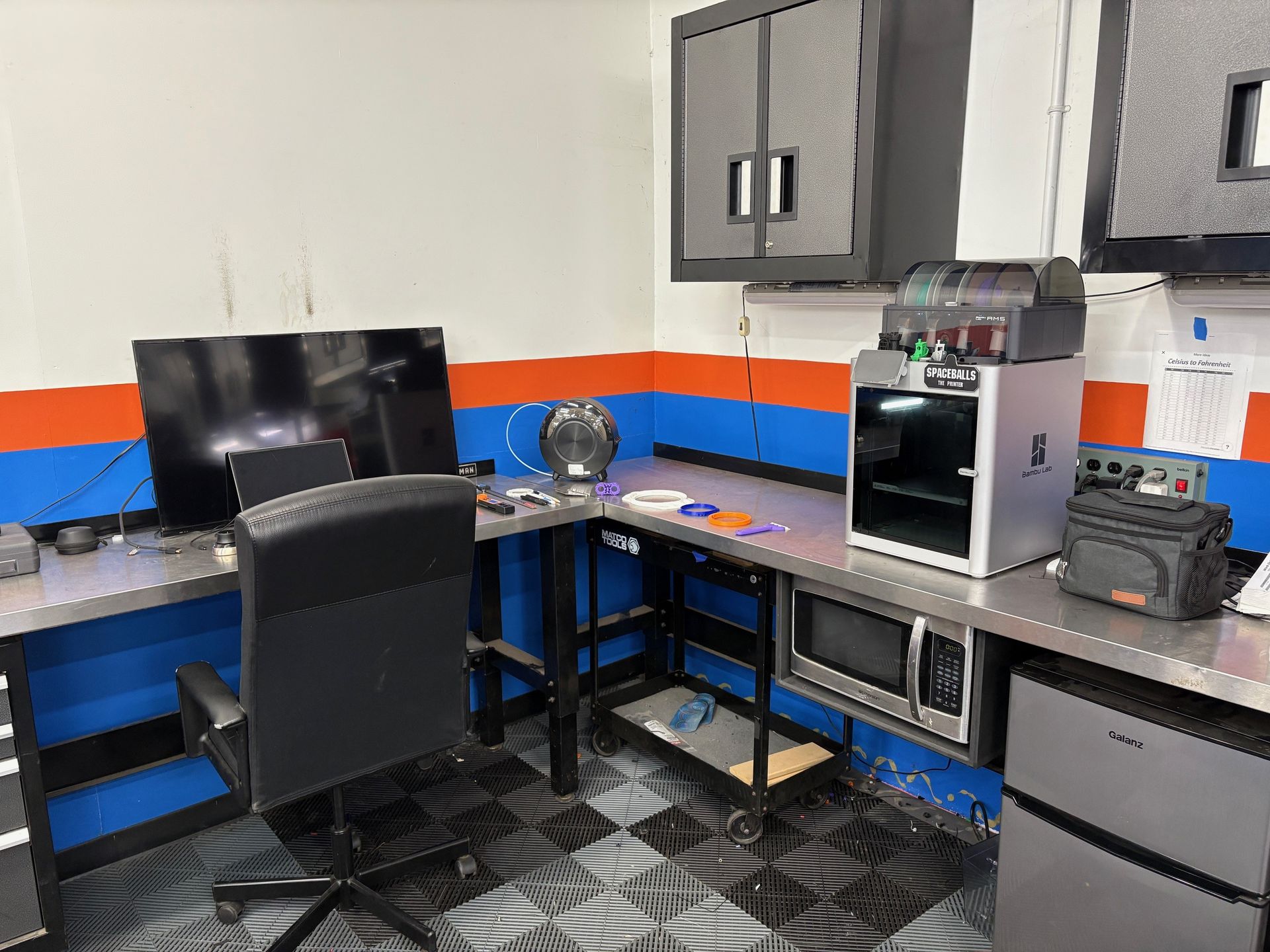 A room with a desk , chair , computer and microwave.