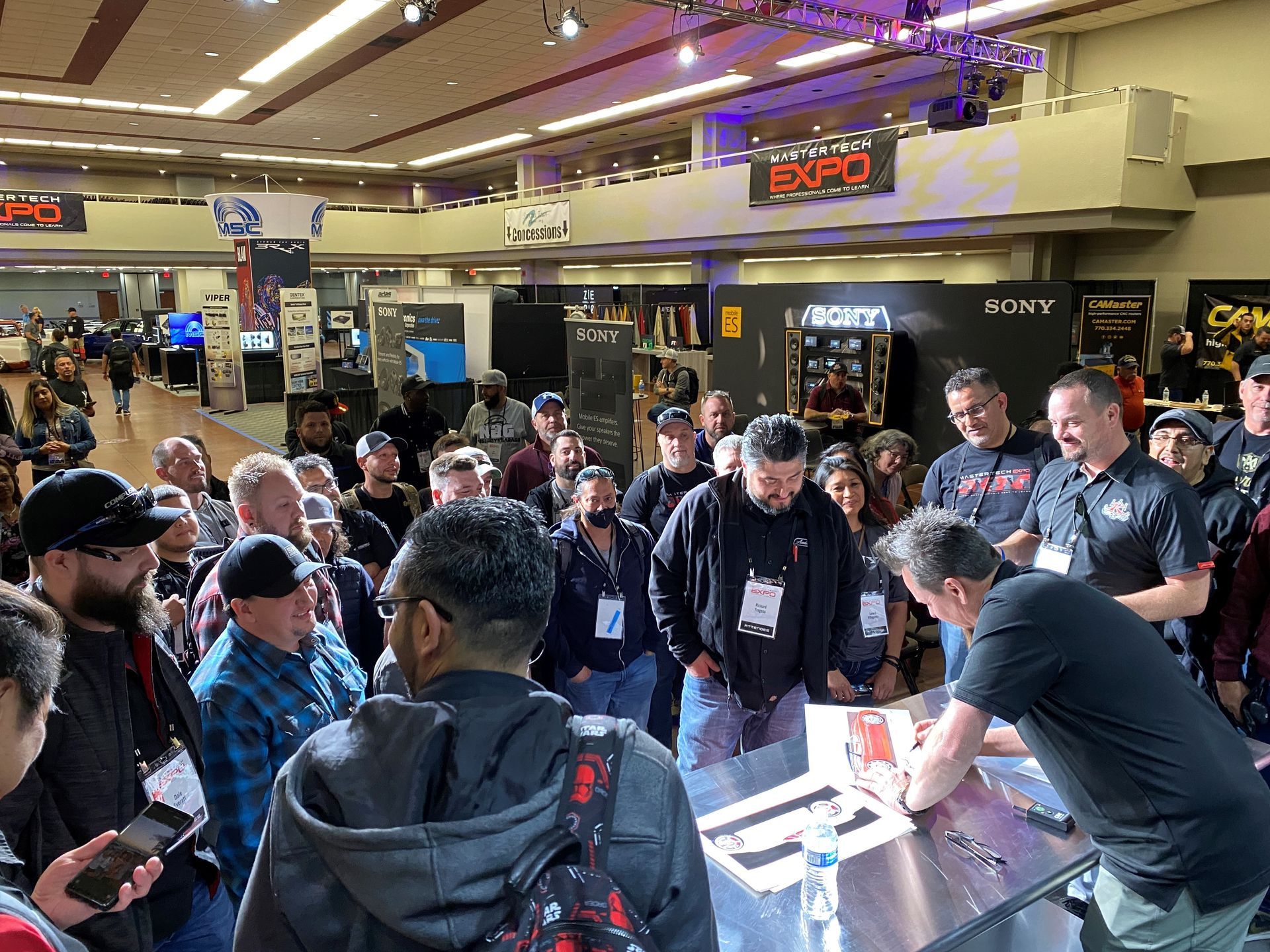 A group of people are standing around a table in a room with a guest signing autographs.