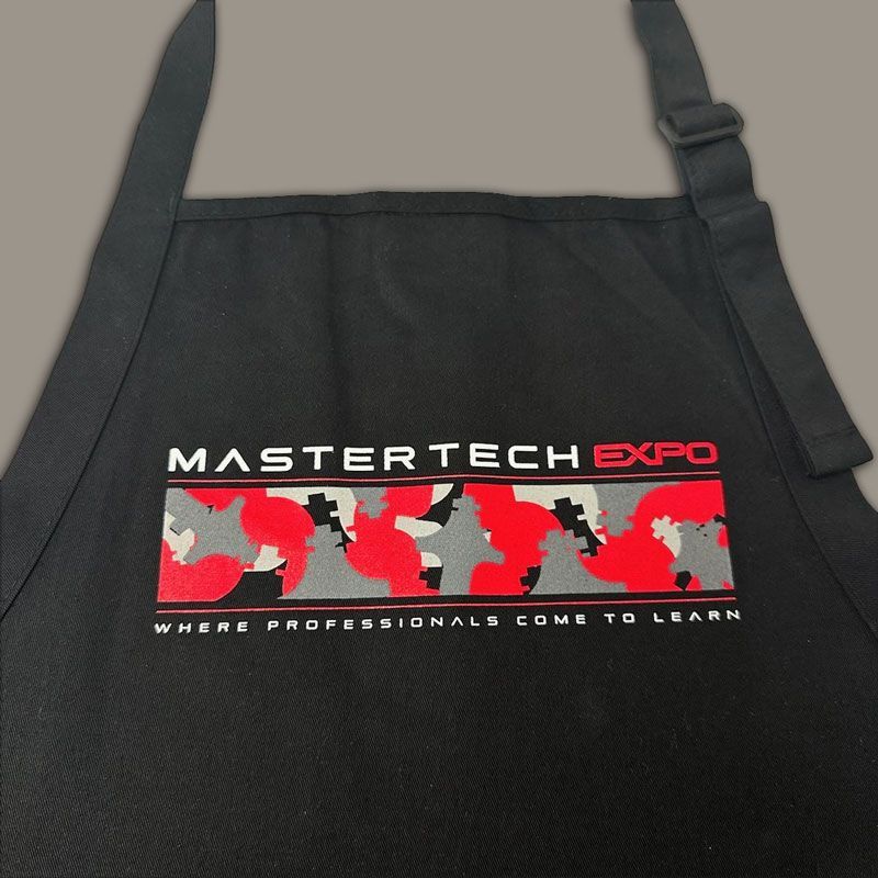 Woman wearing a black apron with a logo, standing near metal drawers, indoors.