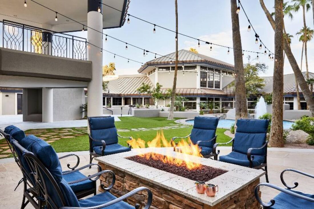 Outdoor patio with a fire pit surrounded by blue chairs, buildings, palm trees, and string lights.