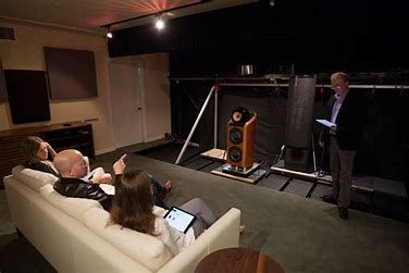 A group of people are sitting on a couch in a living room while a man stands in front of a speaker.