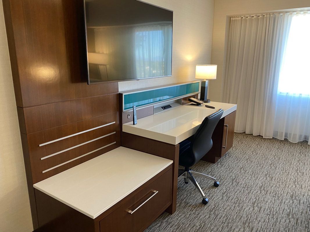 Hotel room desk with a mounted TV, lamp, and chair, next to a window with sheer curtains.