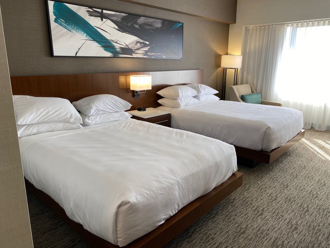 Two queen beds in a hotel room, with white bedding, nightstand, art, and a sunny window.