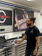 Man with topknot looking at a screen displaying a 3D model, standing near a laptop and workbench.