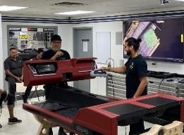 Three men inspecting a red car dashboard in a workshop, another dashboard on a table.