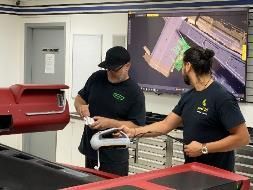 Two men inspecting a car part with a scanner in a workshop, a screen displaying a 3D model.
