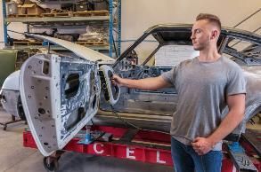 Man in gray shirt inspects a car door in a garage. Car is on a lift. Shelves and tools in the background.