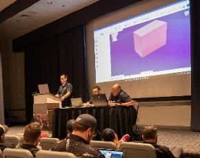 A presentation in a lecture hall. Three people at a table, one at a podium. A screen displays a 3D model.