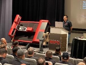 Man presenting at a trade show with a large red machine, audience seated.