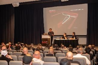 Audience at presentation, speaker at podium, two people seated, projected image on screen.