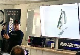 Man presenting a tech item, pointing at a screen with a similar item's image. In a room with posters and tech on a table.