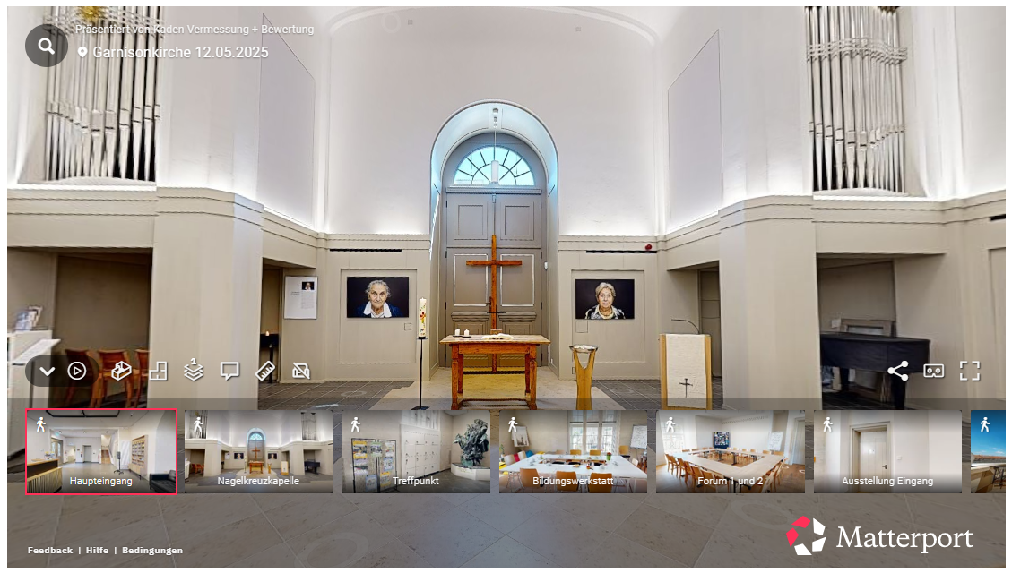 Innenraum einer Kirche. Helle Wände mit einem gewölbten Eingang. Holzkreuz und Altar. Zwei gerahmte Bilder hängen.