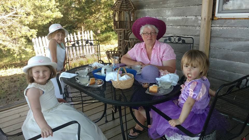 Group of people, including children, seated at a table on a porch, enjoying tea and treats.