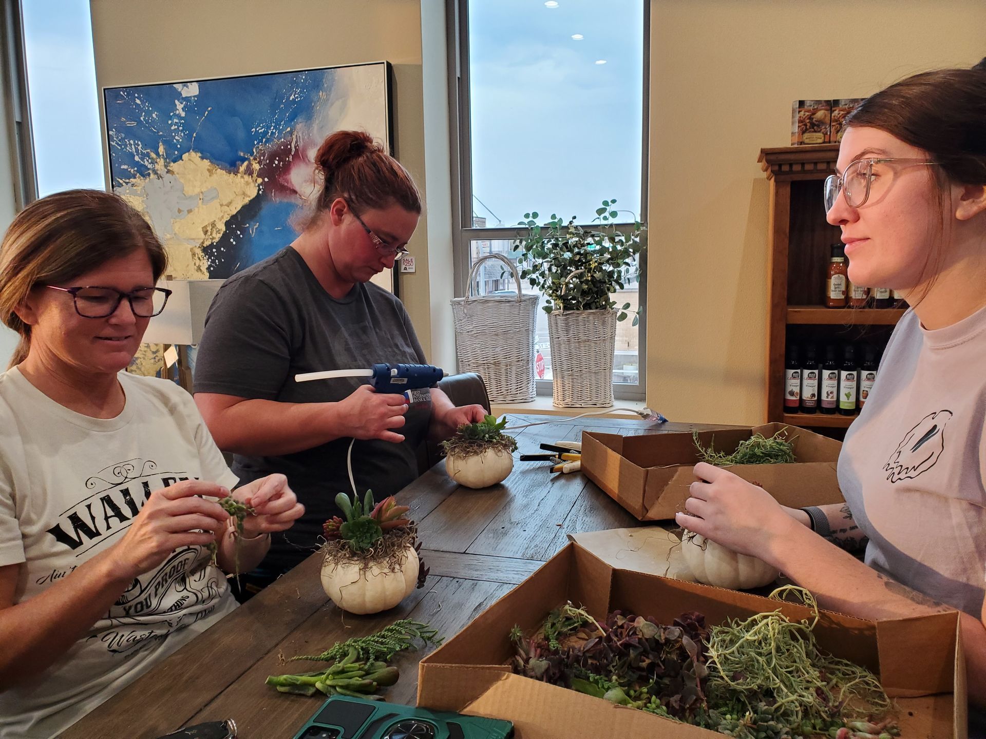 Three people crafting with succulents and pumpkins at a table. One uses a glue gun. Indoors, natural light.