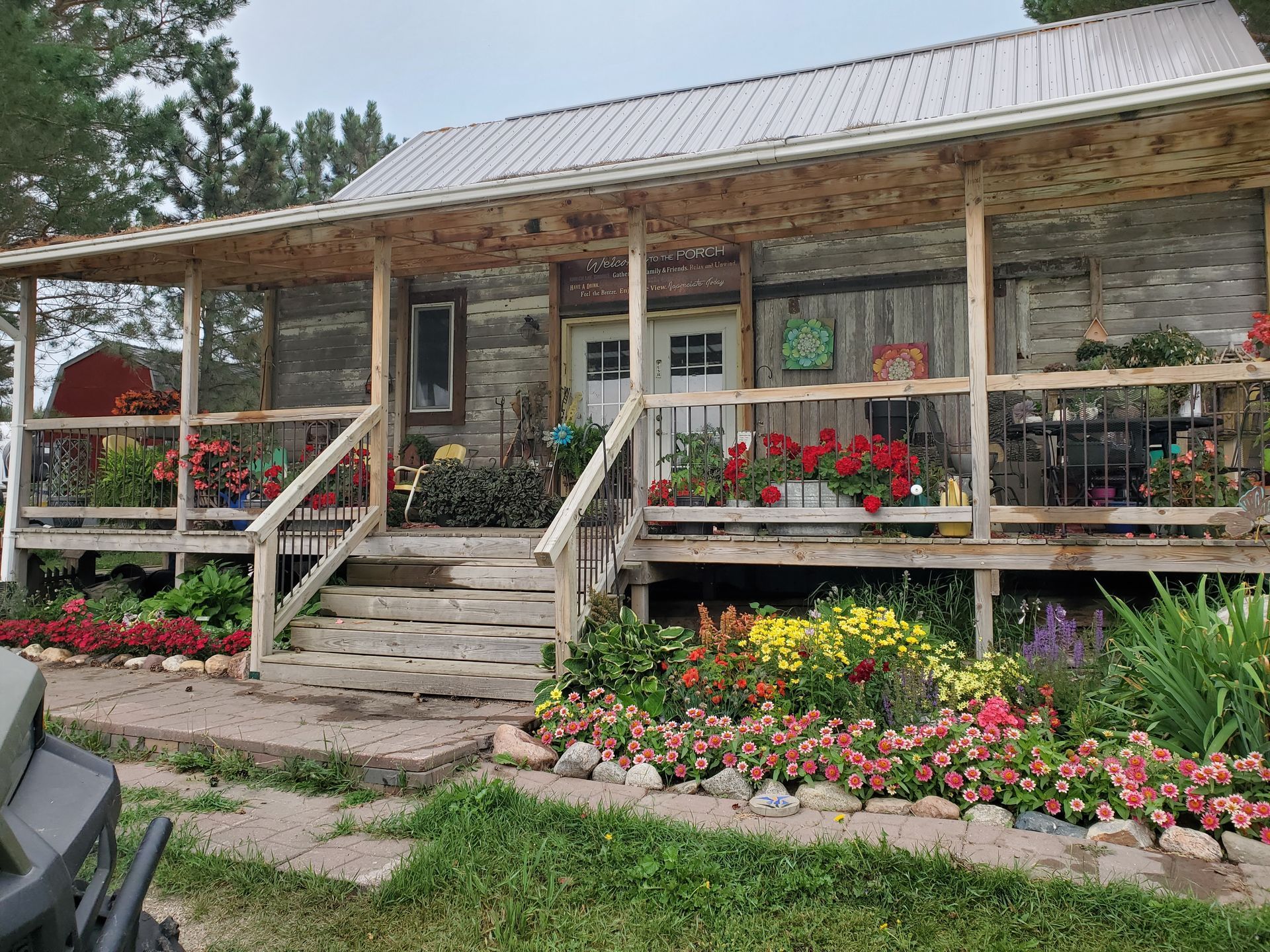 Log cabin with a wraparound porch, surrounded by flower gardens and steps leading up to the front door.